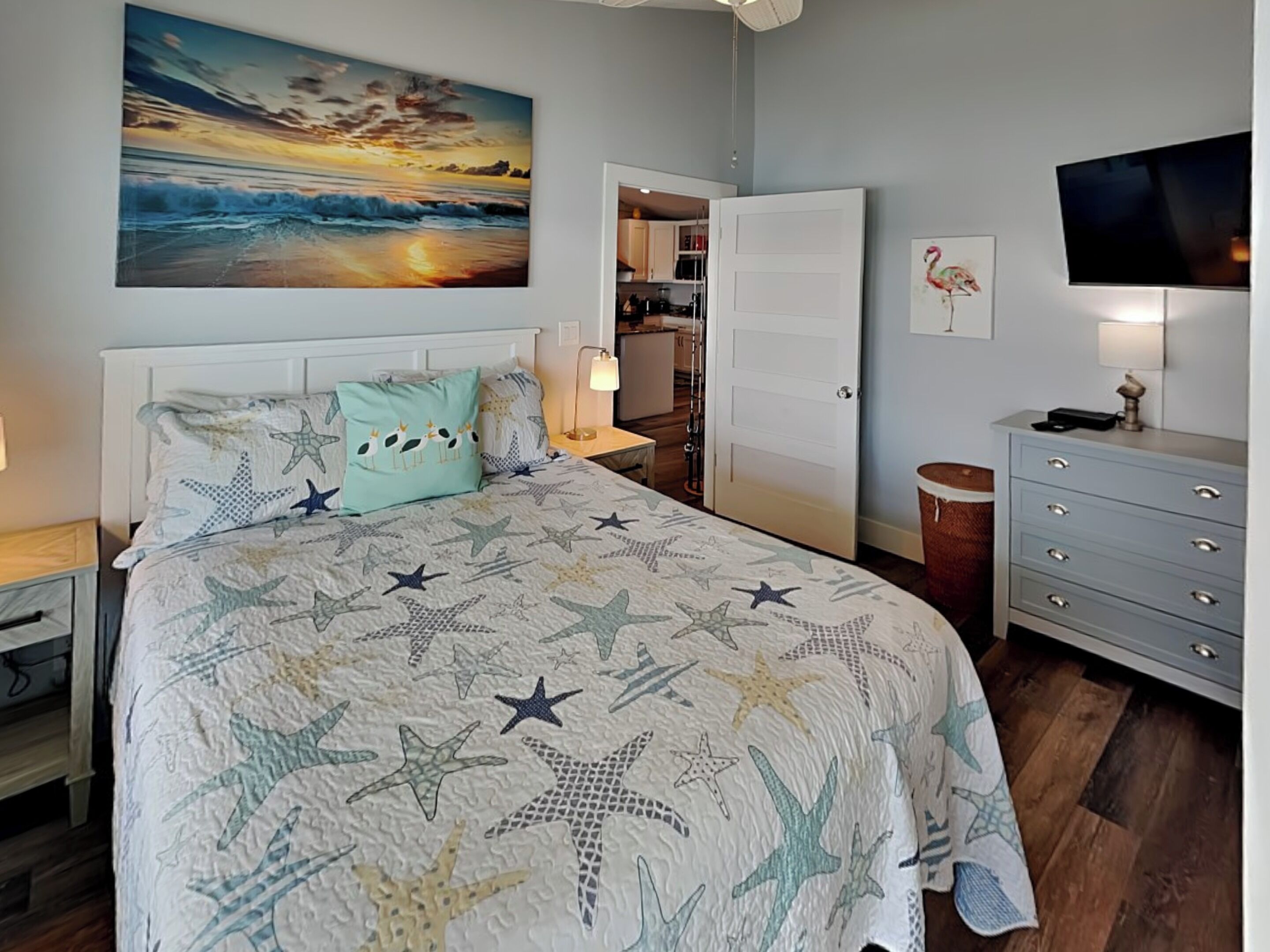 A bedroom with a bed featuring a starfish and seahorse pattern, a painting of a sunset, a television, and a wooden floor.