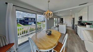 A modern kitchen with white cabinets, a dining area with a wooden table and white chairs, and a view of a blue house through large windows. - 275 Blue Bay by Distinctive Beach Rentals (Fort Myers Beach)