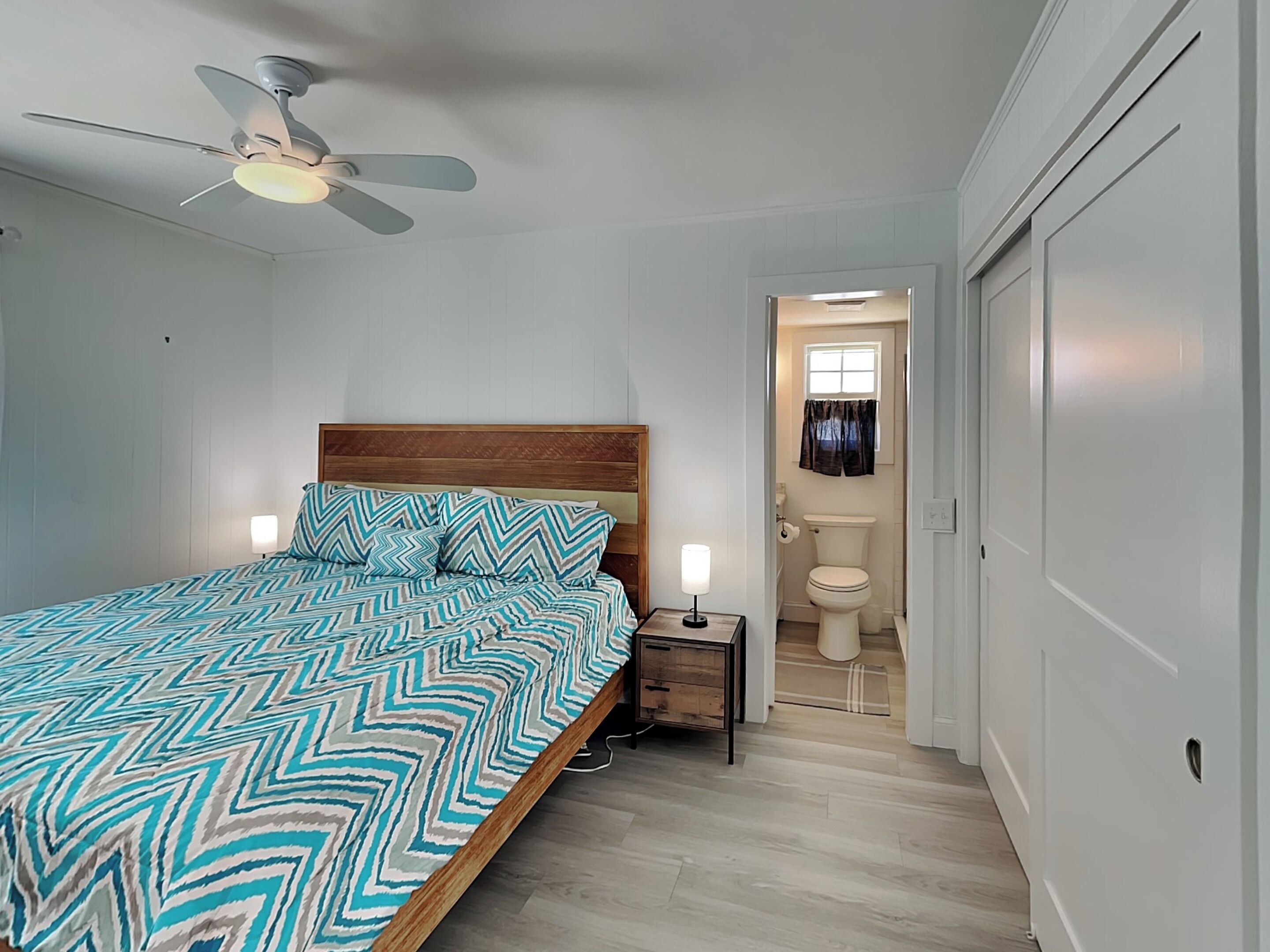Une chambre à coucher comprenant un lit, une table de chevet, un ventilateur de plafond et donnant sur la salle de bain.