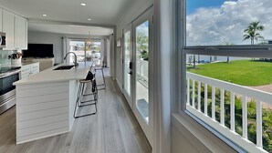 A modern kitchen with a large island, a dining area, and a view of a green lawn and palm trees through large windows.