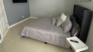 Un dormitorio con una cama, una mesita de noche con un control remoto, un televisor montado en la pared y una lámpara en el techo.