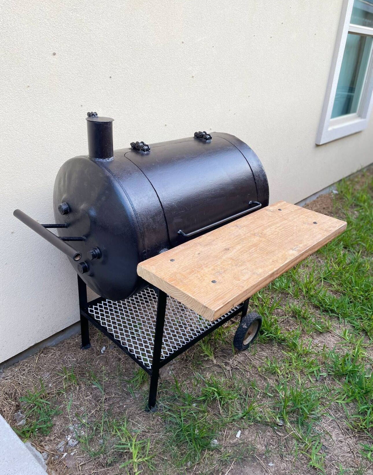 Una griglia barbecue portatile con un tavolo di legno su ruote, posizionata sull'erba vicino a un edificio.