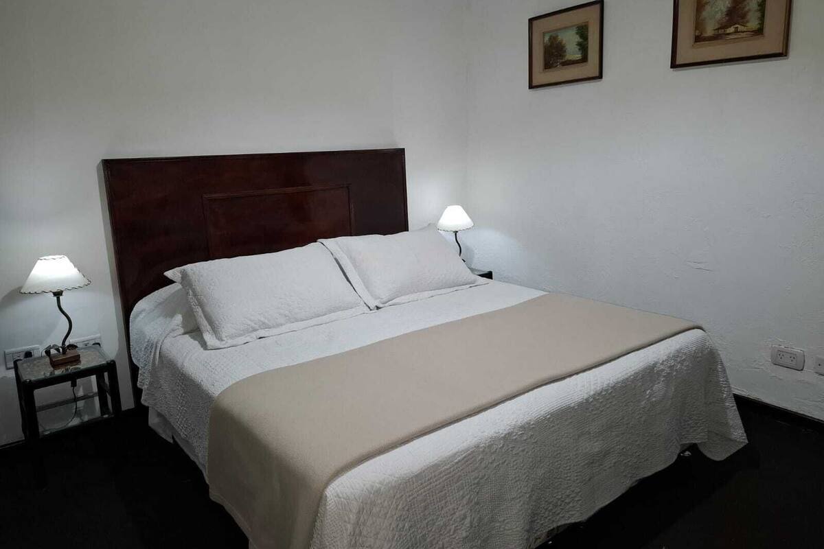 Un llit ben fet amb capçalera de fusta, dues làmpades de nit i dues fotografies emmarcades a la paret.