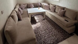 A living room with a beige sectional sofa, a white coffee table, a shaggy rug, a hanging light fixture, and a window with blinds. - marrakesh apartment (Marrakech)