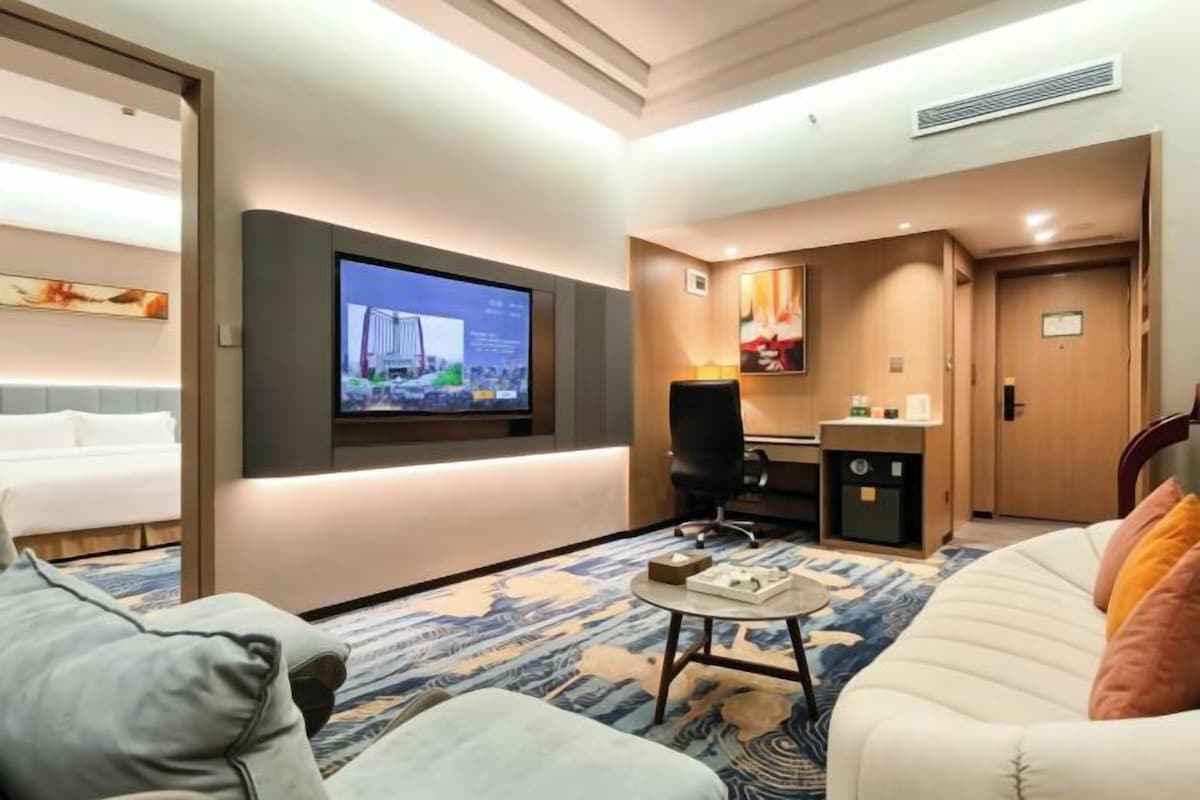 Quarto de hotel com cama, sofá, mesa de centro, TV fixada na parede e escrivaninha com cadeira.