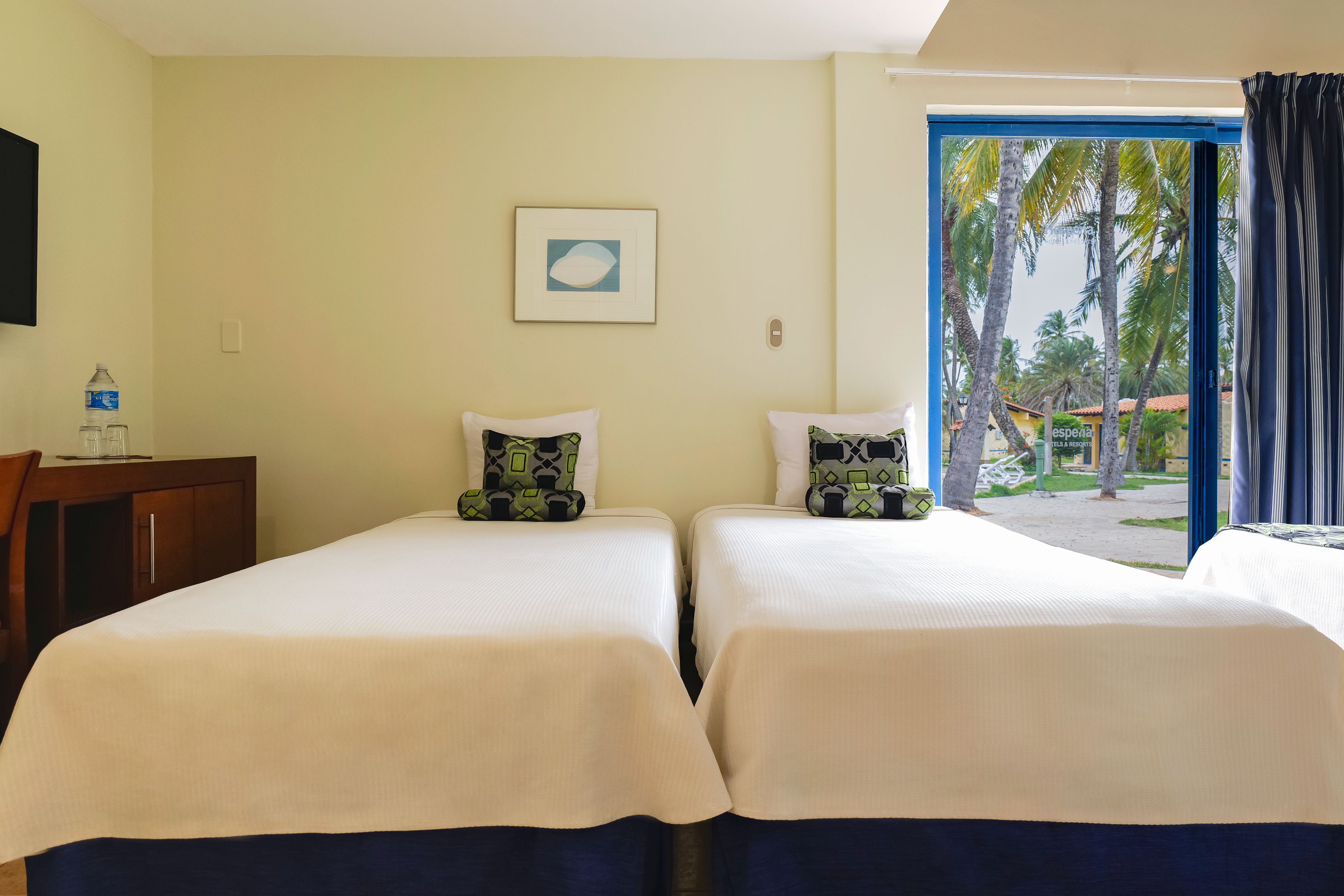 Habitación de hotel con dos camas, una botella de agua sobre una mesita de noche, un cuadro en la pared y vistas a palmeras a través de la ventana.