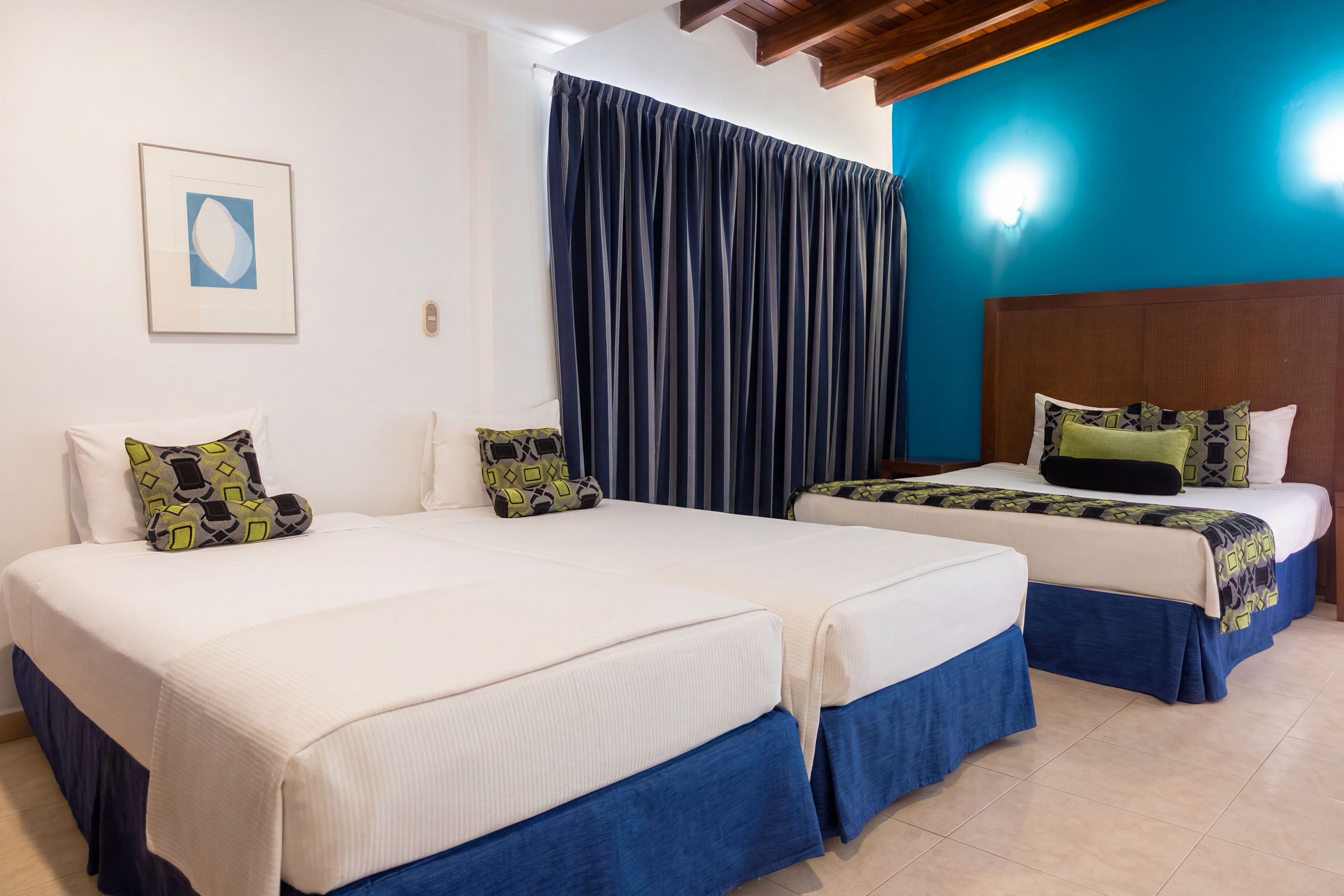 A hotel room with two beds, a large window with curtains, a wooden ceiling, and a framed picture on the wall.