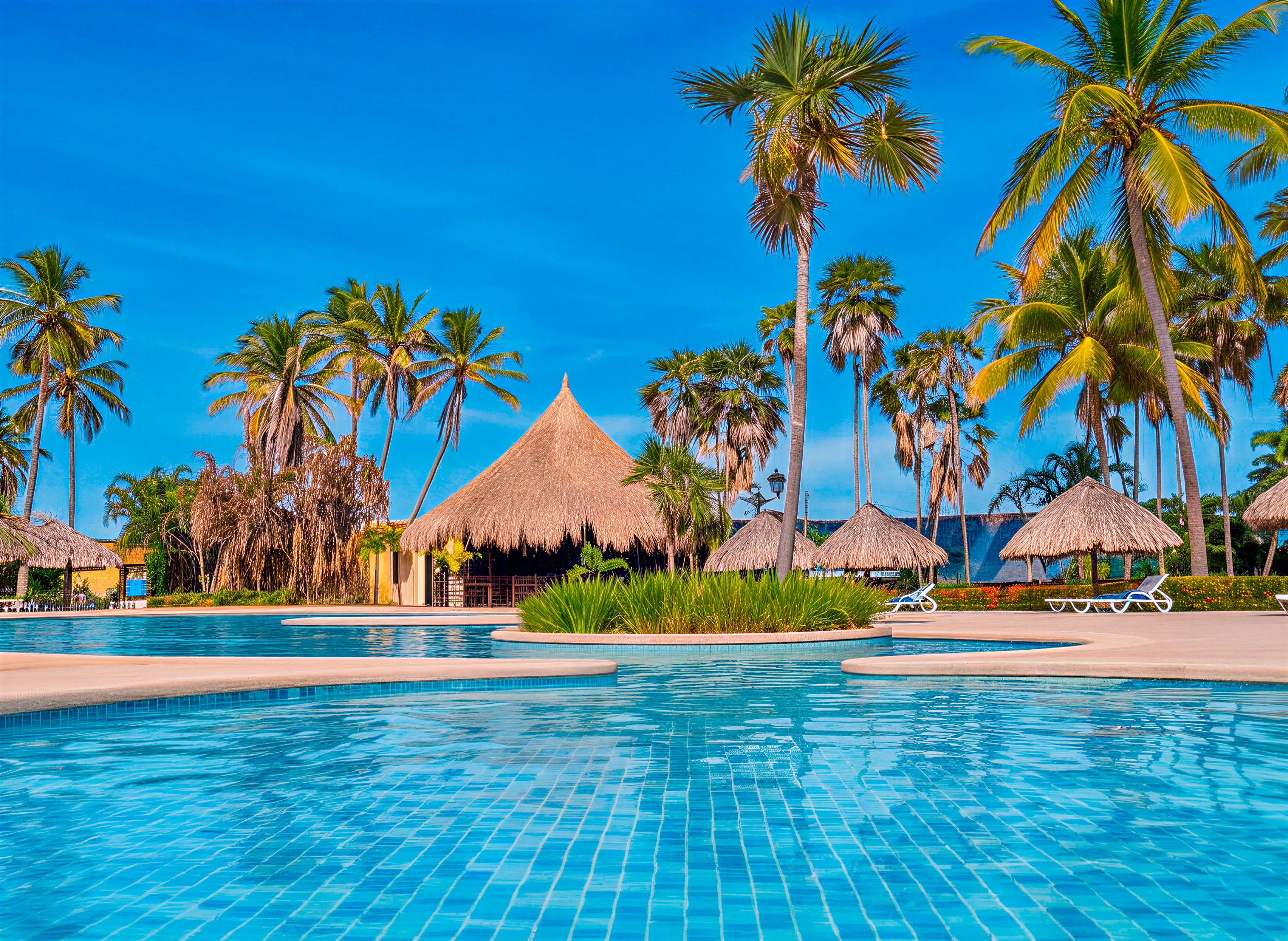 Een zwembad in een resort met palmbomen en hutten met rieten daken.