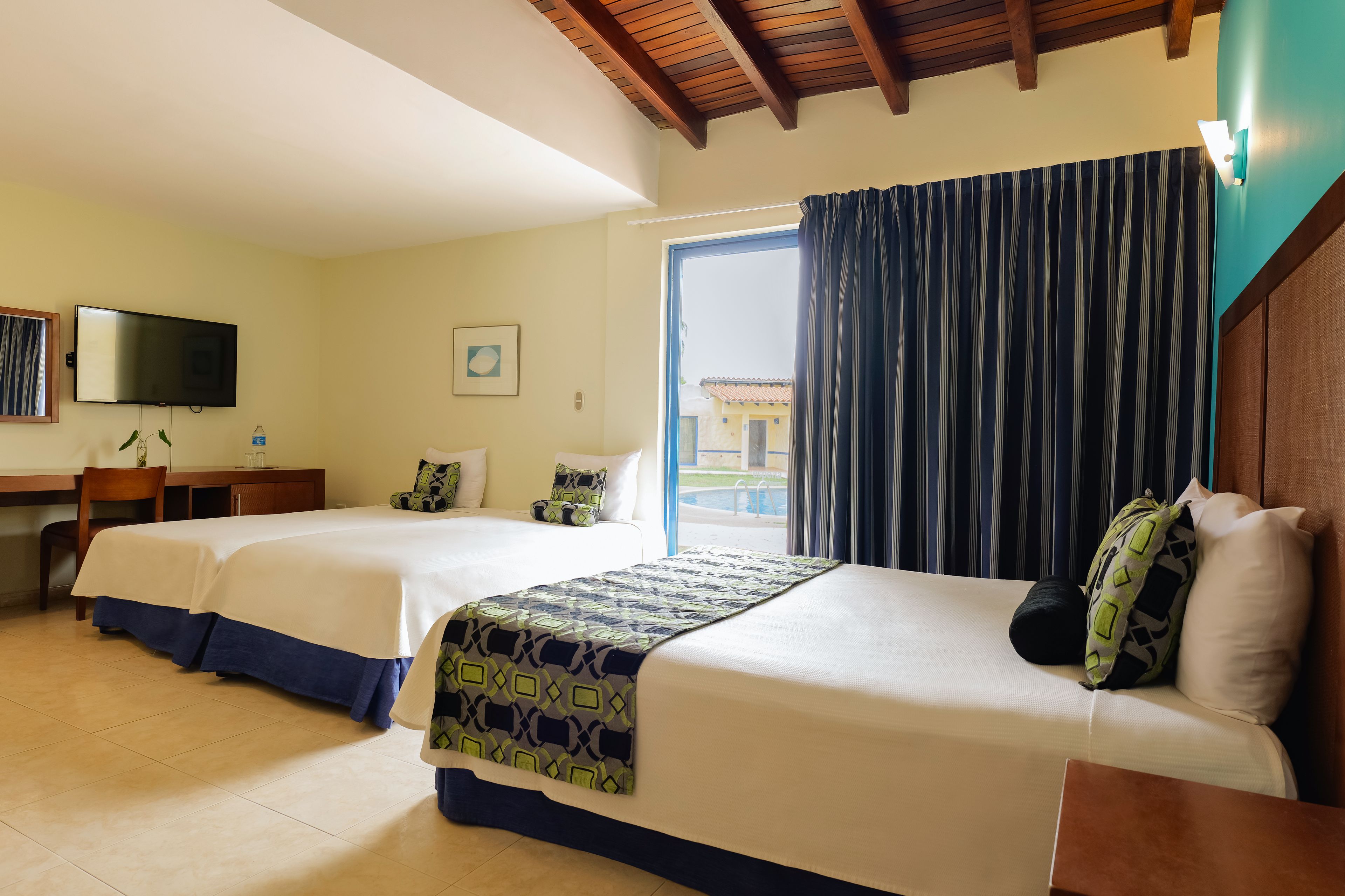 Habitación de hotel con dos camas, un escritorio, un televisor y un ventanal con cortinas.