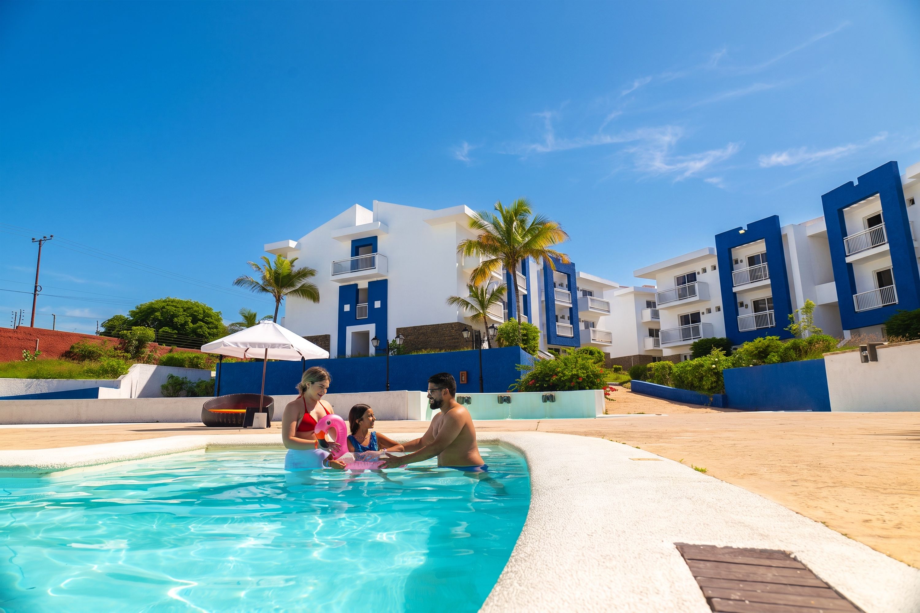 A swimming pool area with a clear blue sky, a couple and a child in the pool, and modern buildings with balconies in the background.