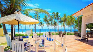 Un patio frente a la playa con sillas y mesas blancas, un paraguas amarillo y vista al océano y palmeras.
