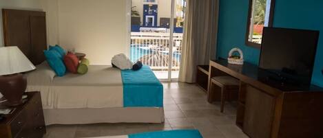 Habitación de hotel con cama, escritorio, televisión y vista a una piscina a través de una puerta corrediza.