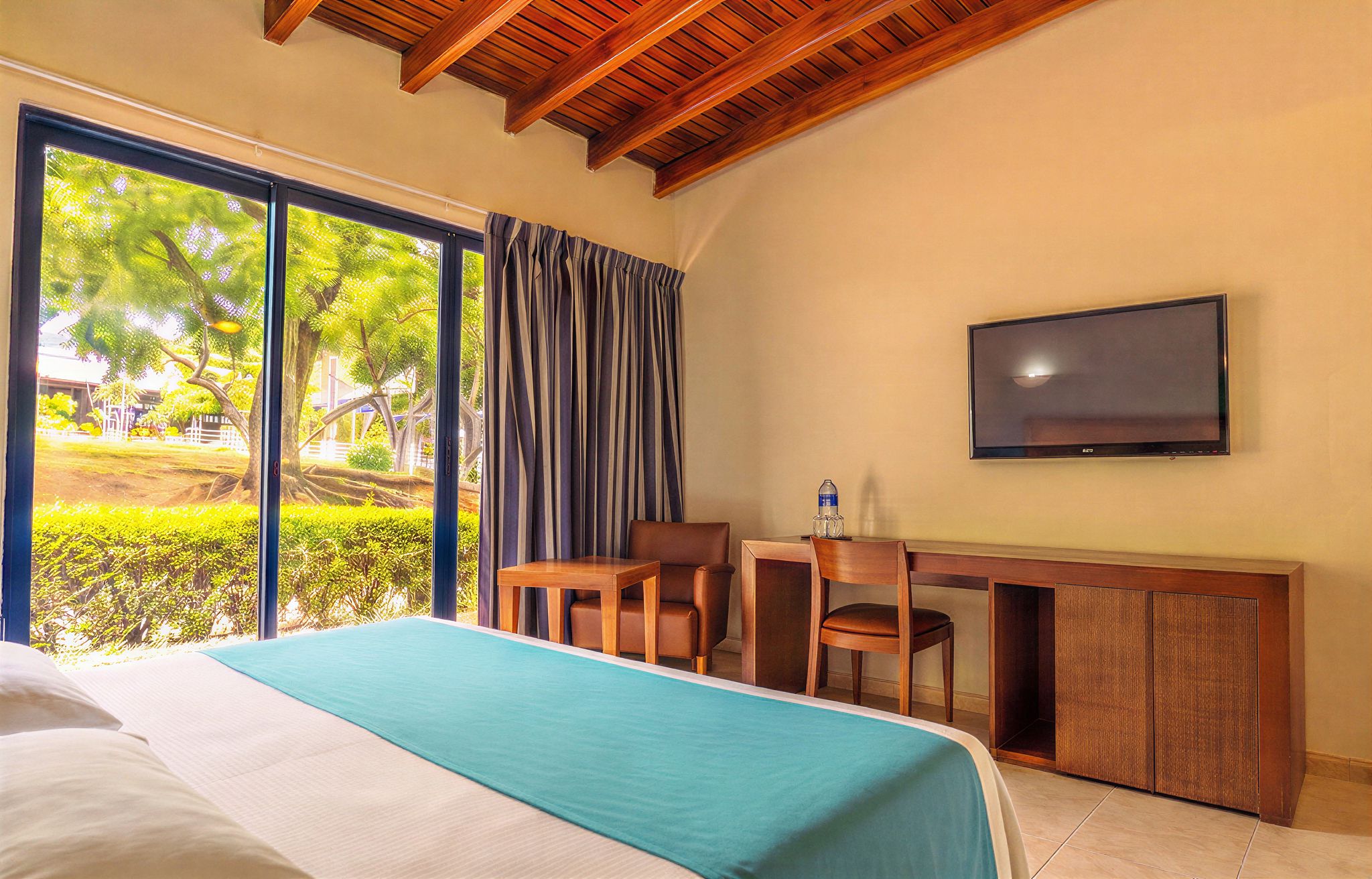 A hotel room with a bed, a desk with chairs, a television, and a large window with curtains.