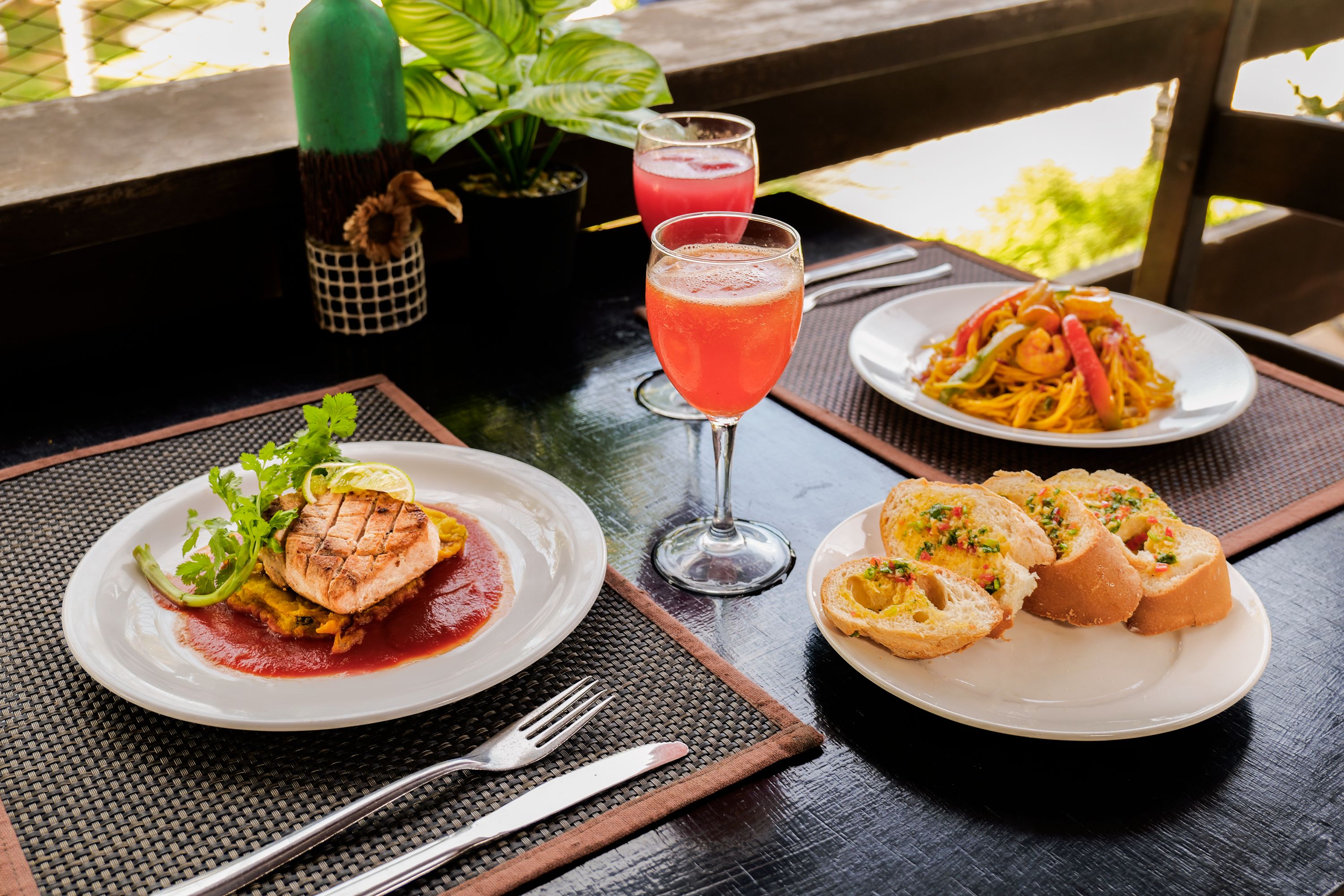 A table with a meal consisting of grilled fish, a red drink, and a plate of noodles.