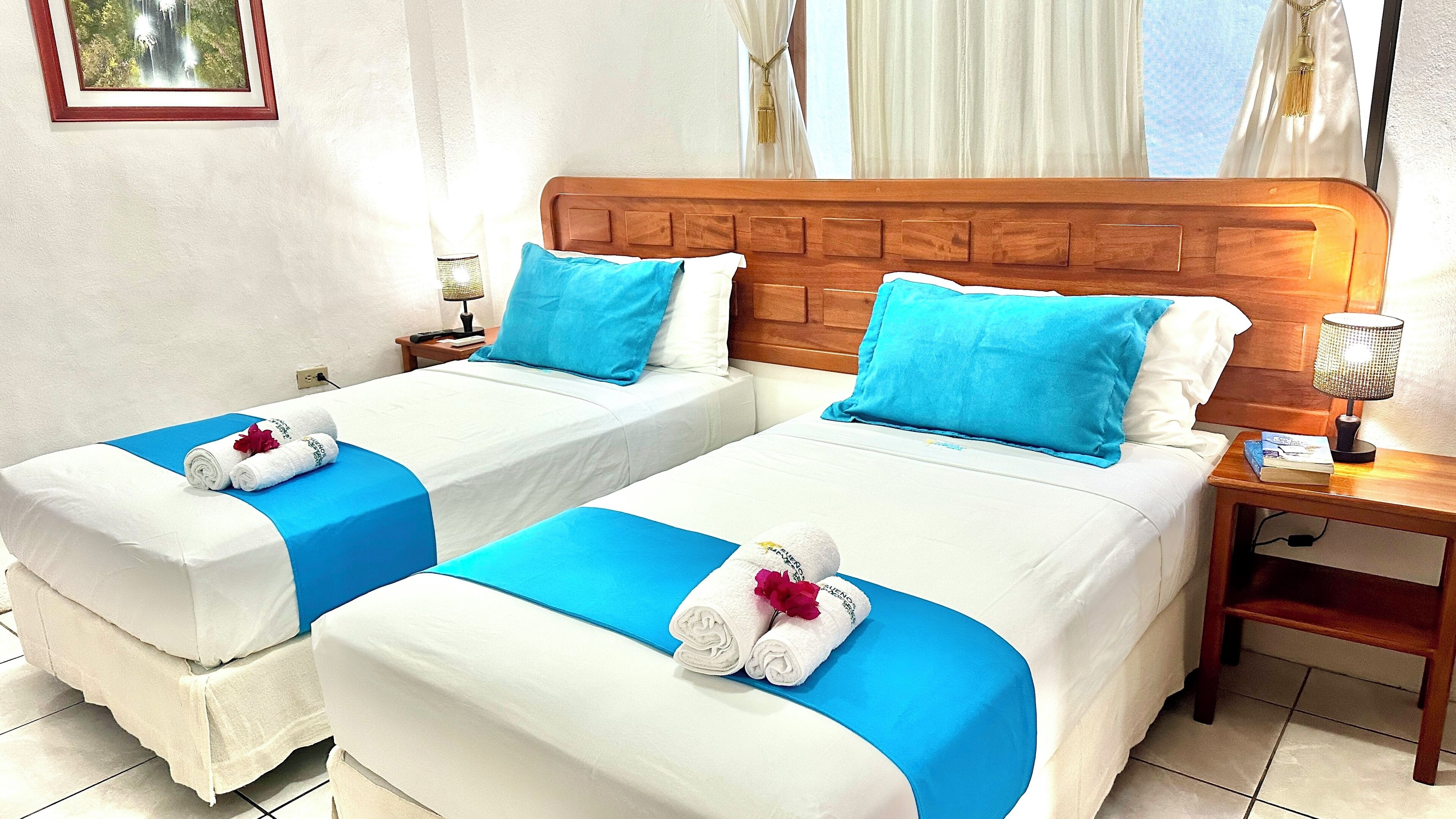 Two single beds with blue bedspreads and white linens, a wooden headboard, a nightstand with a lamp, and a framed picture on the wall.