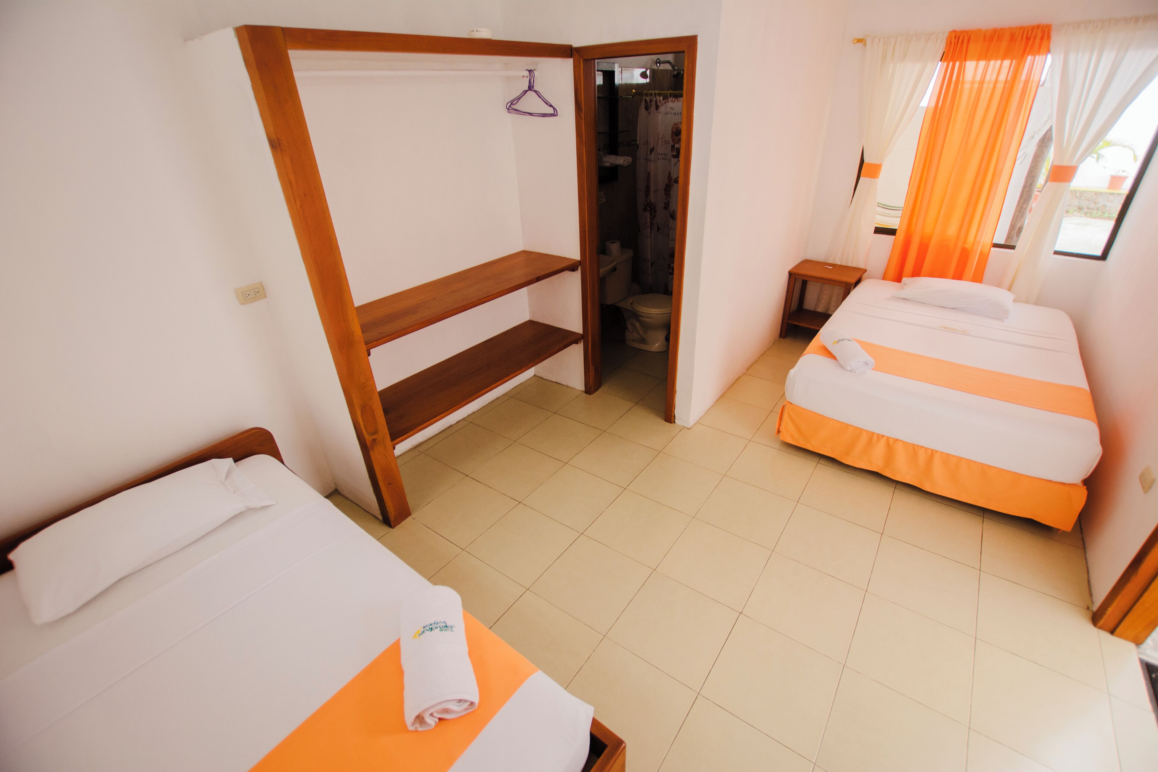 A hotel room with two beds, a wooden wardrobe, a bathroom with a shower curtain, and a small table.