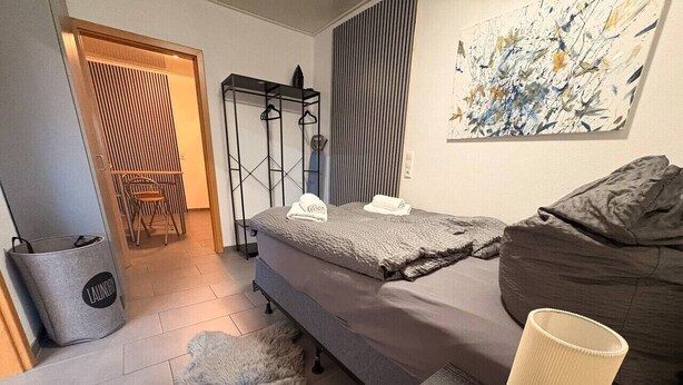 A compact bedroom with a bed, a small table, a lamp, a fur rug, a chair, a wardrobe, and a painting on the wall. - Studio One - 5 min to Grafenwöhr (Pressath)