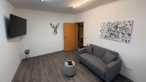 A modern living room with a grey sofa, a flat-screen TV mounted on the wall, a wooden door, and a colorful abstract painting. - Studio One - 5 min to Grafenwöhr (Pressath)