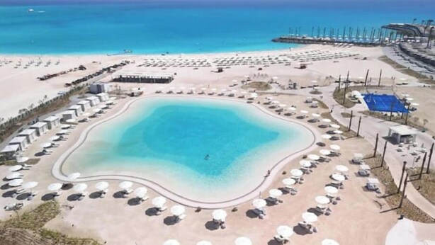 An aerial view of a beach resort with a central pool, sun loungers, and a pier extending into the sea.