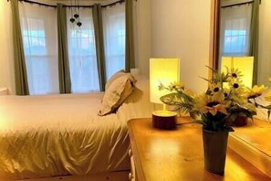 A hotel room with a bed, a bedside table with a lamp and a vase of flowers, and a window with curtains. - Modern Shepherd's Lodge with stunning 360 Views, Fenced Pet Area and Pool (Buncombe County)