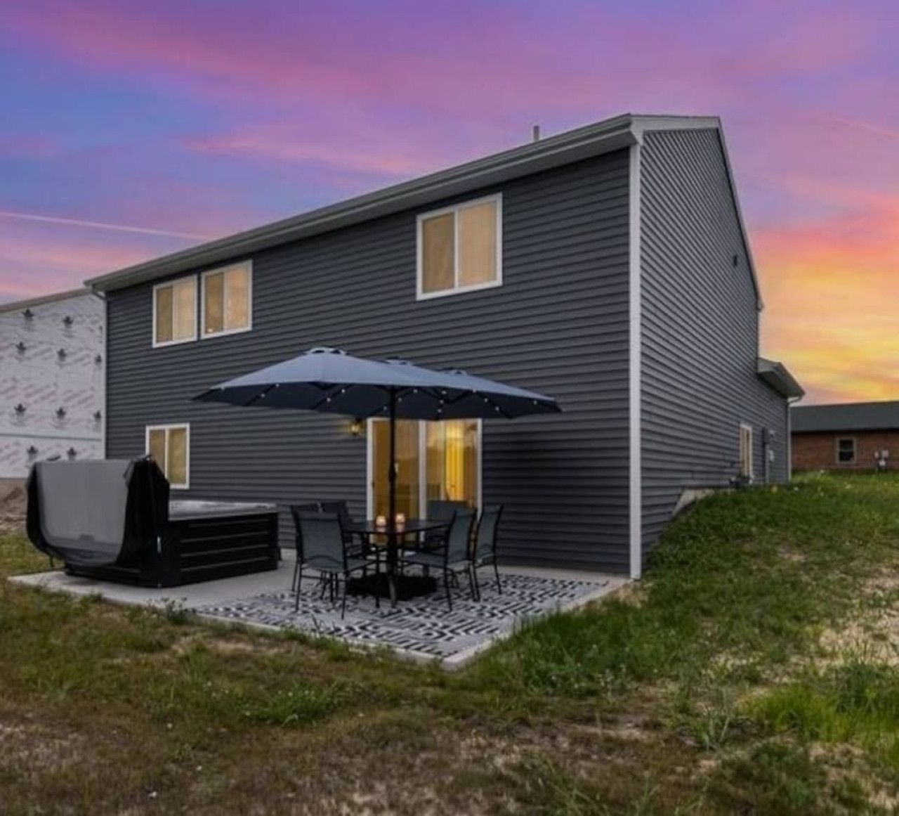 A modern two-story house with a patio area featuring a grill, outdoor seating, and a large umbrella.