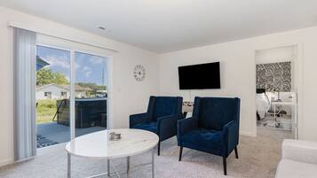 A modern living room with a white marble table, two dark blue armchairs, a flat-screen TV mounted on the wall, and a sliding glass door leading to an outdoor area.