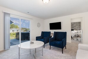 Een moderne woonkamer met een witte marmeren tafel, twee donkerblauwe fauteuils, een flatscreen-tv aan de muur en een glazen schuifdeur naar een buitenruimte.