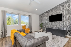 Un salon moderne avec un canapé gris, des fauteuils jaunes, une table basse blanche et un grand téléviseur fixé sur un mur à texture.