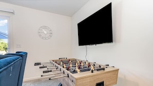 A room with a foosball table, a blue sofa, a wall clock, and a mounted TV. - Brand New House with Hot Tub! Gameroom! Workspace! (Michigan City)