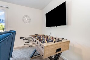 Une pièce avec une table de baby-foot, un canapé bleu, une horloge murale et un téléviseur fixé au mur.