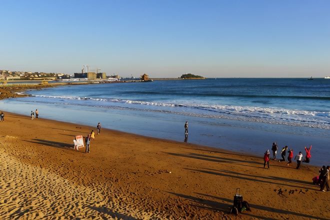 A beach with people, a boat, and industrial structures in the background.
