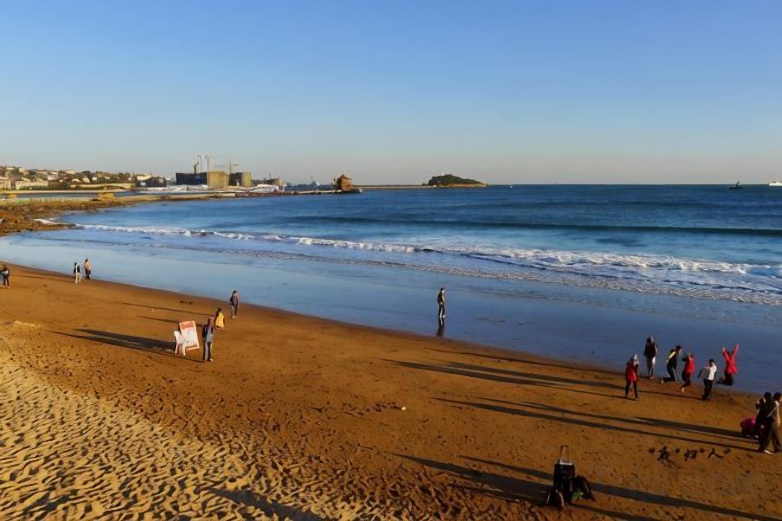 Une plage où l’on voit des gens se promener le long du rivage, un ciel dégagé et des bâtiments qui se dressent au loin.
