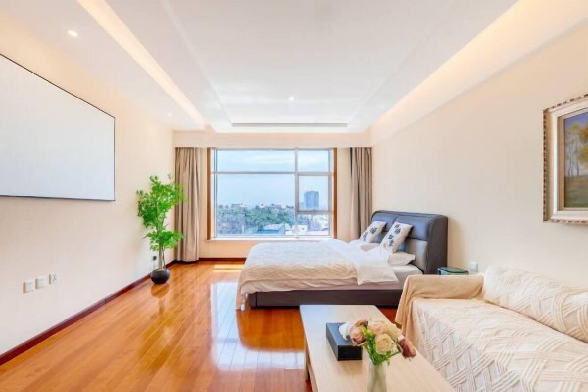 A modern hotel room with a large bed, a sofa, a desk, and a large window with a city view.