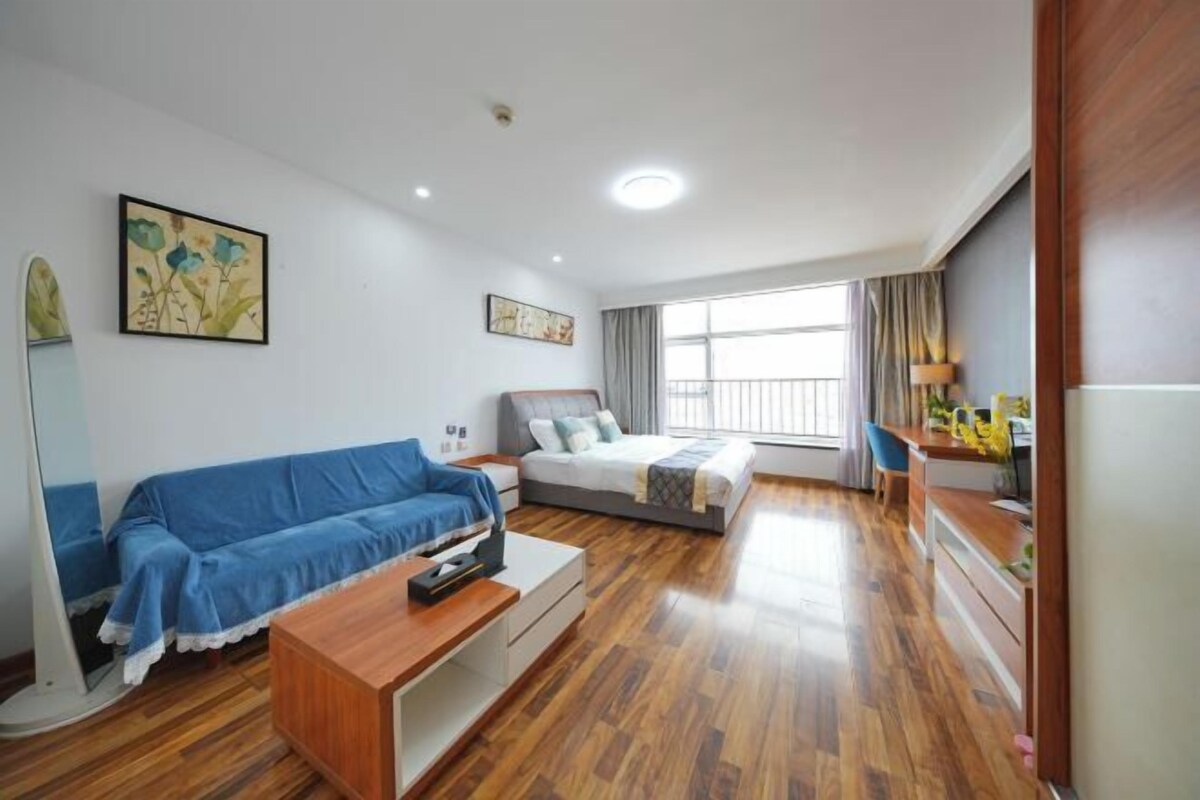 A modern hotel room with a wooden floor, a blue sofa, a bed with a grey headboard, a desk with a lamp, and a window with curtains.