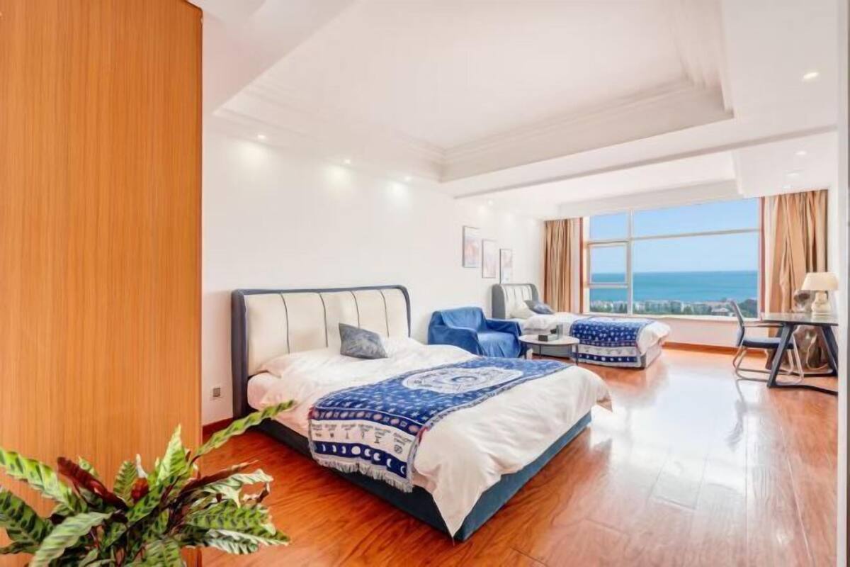 A hotel room with a bed, a desk, a chair, a view of the sea, and a plant.