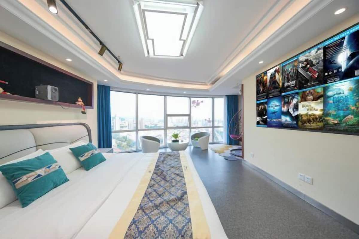 A hotel room with a large bed, a long patterned rug, a wall with movie posters, and a large window with a city view.