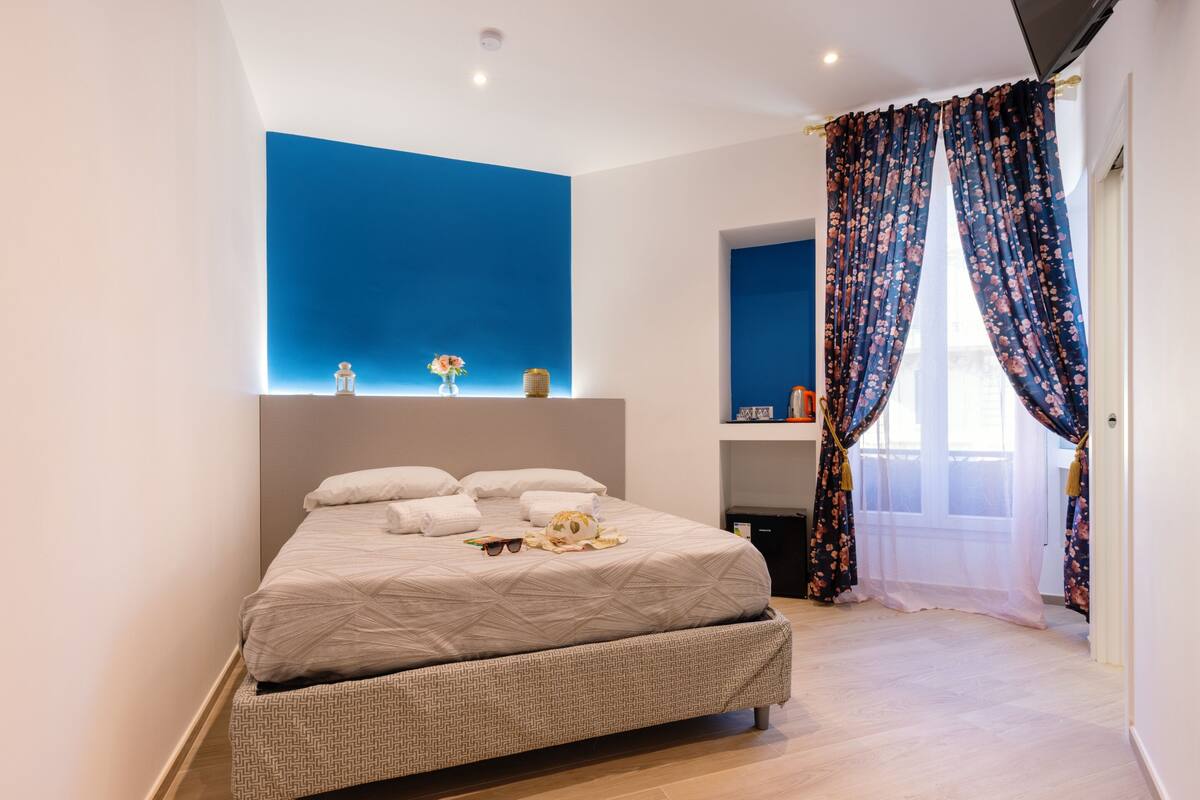 A modern bedroom with a bed, a bedside table, a small shelf, and a window with curtains.