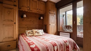 A bedroom with a wooden headboard, a bed with a red and white patterned bedspread, two pillows with animal designs, and a window with a view of a balcony and trees.
