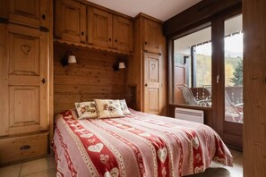 A bedroom with a wooden headboard, a bed with a red and white patterned bedspread, two pillows with animal designs, and a window with a view of a balcony and trees.