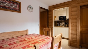 A room with a bed, a table, and chairs. There is a wooden door leading to a kitchen area with a microwave and a coffee maker.