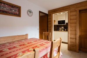 Quarto com uma cama, uma mesa e cadeiras. Há uma porta de madeira que dá para uma área de cozinha com micro-ondas e cafeteira.