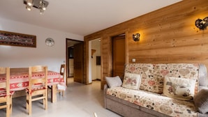 A cozy living room with a wooden sofa, a dining table with chairs, and a wooden wall with a heart-shaped light fixture.