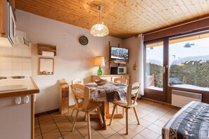 A cozy room with a wooden ceiling, a dining table with chairs, a TV, a microwave, and a large window with a view of trees. - 250m from ski slopes, Ground floor, terrace, tv, ski locker, 28m², La Clusaz (La Clusaz)