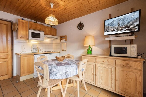 A cozy kitchen with wooden cabinets, a dining table with chairs, a hanging light fixture, a television, and a microwave.