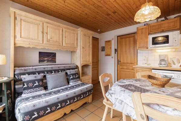 A cozy room with a sofa bed, wooden cabinets, a dining table, and a kitchen area with a microwave.