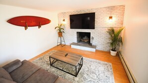 A living room with a fireplace, a mounted surfboard, and a television. - 5 STAR Coastal Farmhouse- Dog Friendly- 3BR in Westerly! Minutes to Beach & Town (Westerly)