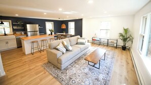 A modern kitchen with a central island, bar stools, and a sofa in a living area with a patterned rug. - 5 STAR Coastal Farmhouse- Dog Friendly- 3BR in Westerly! Minutes to Beach & Town (Westerly)
