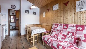 A cozy room with a wooden ceiling, a dining table with chairs, a sofa with a patterned cover, and a wall clock.