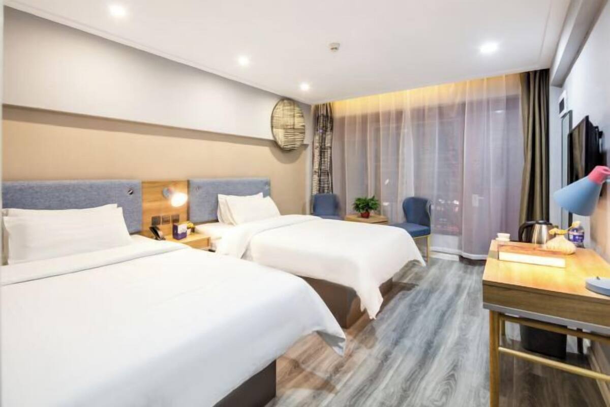 A hotel room with two beds, a desk, a chair, a plant, and a large window with curtains.