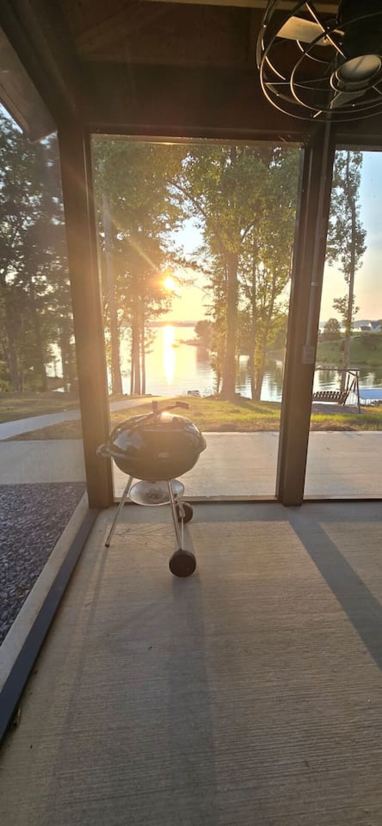 A view from a room with a glass door, overlooking a body of water and trees, with a barbecue grill in the foreground.